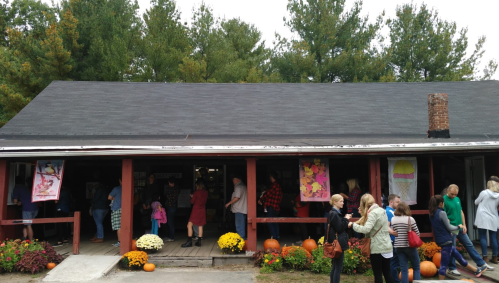A busy outdoor market with people browsing, pumpkins, and colorful flowers in front of a rustic building.