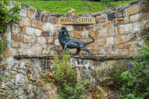 A stone wall with a plaque reading "Berkeley Castle 1865" and a bronze lion statue in front of it.