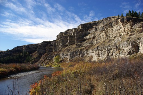 A scenic view of rocky cliffs beside a winding river, surrounded by autumn foliage under a blue sky.
