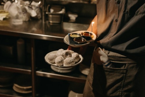 A server holds two plates: one with powdered donuts and the other with dipping sauces, in a dimly lit kitchen.