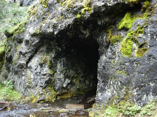 A moss-covered rock formation with a dark cave entrance, surrounded by greenery and a small stream.