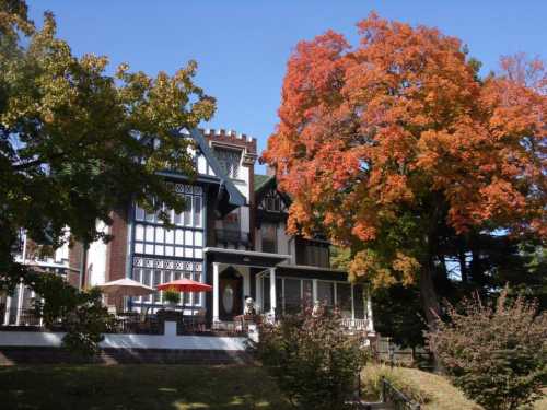 A large, historic house with a colorful autumn tree in front, showcasing vibrant red and orange leaves against a clear blue sky.