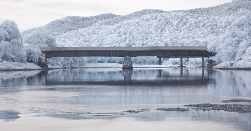 A serene landscape featuring a covered bridge over a calm river, surrounded by trees and mountains in a soft, muted palette.