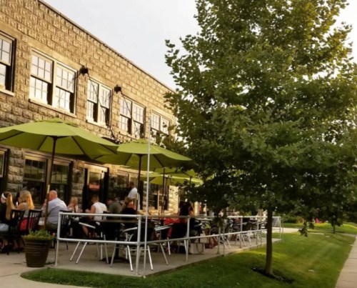 Outdoor dining area with green umbrellas, people seated at tables, and a tree nearby in a sunny setting.