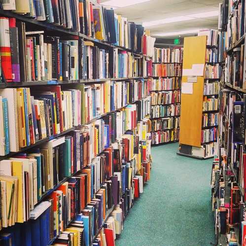 A narrow library aisle lined with tall bookshelves filled with various books, leading to a corner with a wooden door.