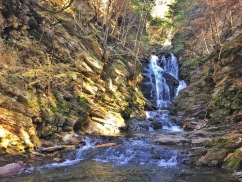 A serene waterfall cascades over rocky terrain, surrounded by lush greenery and sunlight filtering through the trees.