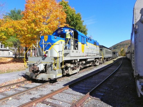A blue and yellow train engine parked on tracks, surrounded by autumn trees with vibrant orange leaves.
