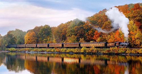 A steam train travels along a river, surrounded by vibrant autumn foliage and reflecting in the water.