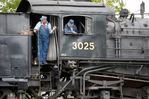 Two train engineers in overalls operate a vintage steam locomotive, with the number 3025 visible on the side.