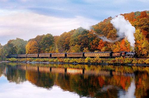 A steam train travels along a river, surrounded by colorful autumn foliage reflecting in the water.