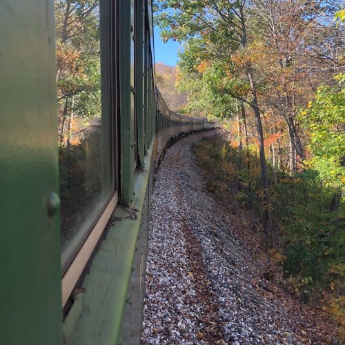 A train curves along a scenic route, surrounded by colorful autumn trees and rocky terrain.