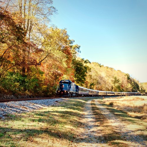A blue train travels along a scenic route surrounded by colorful autumn trees and a clear blue sky.
