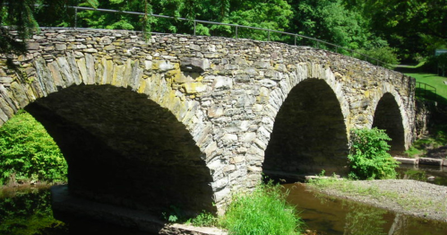 A stone arch bridge with multiple arches spans over a calm stream, surrounded by lush greenery.