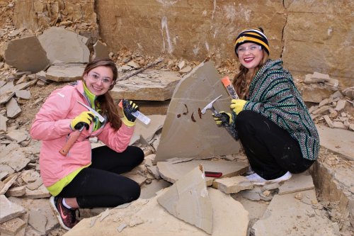 Two women in safety gear are chiseling stone in a rocky area, smiling and working together on a fossil excavation.