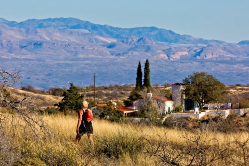 A hiker in a red backpack walks through tall grass with mountains and a small house in the background.