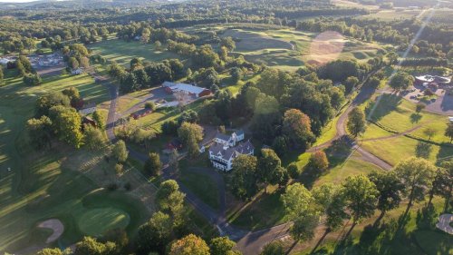 Aerial view of a lush green golf course with trees, a large house, and surrounding farmland under a clear sky.