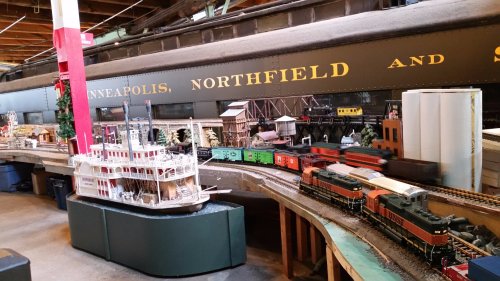 A model train setup featuring various trains, buildings, and a steamboat in a detailed miniature landscape.
