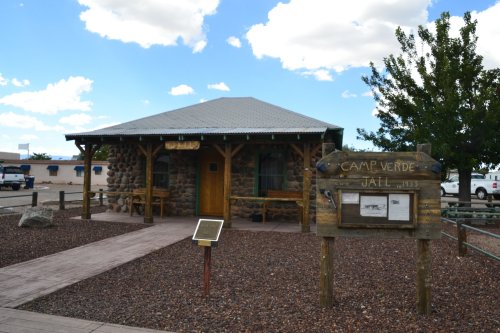 Historic Camp Verde Jail building with stone exterior and wooden accents, surrounded by gravel and trees.