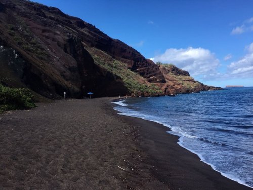 A scenic beach with black sand, rocky cliffs, and calm blue waters under a partly cloudy sky.