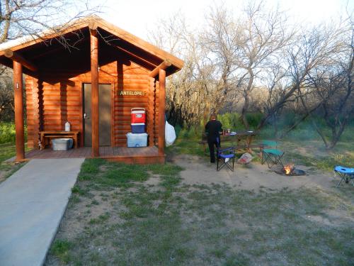 A rustic wooden cabin with a porch, surrounded by trees, and a person cooking near a campfire and chairs.