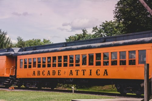 An orange train car with large black lettering reading "Arcade & Attica" against a backdrop of trees and a cloudy sky.