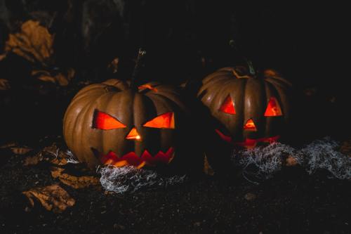 Two carved Halloween pumpkins with glowing faces, surrounded by dry leaves and cobwebs in a dark setting.