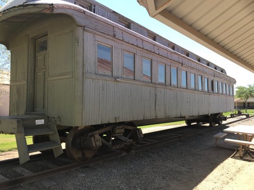A vintage green train car on tracks, with multiple windows and a wooden structure, beside a picnic area.