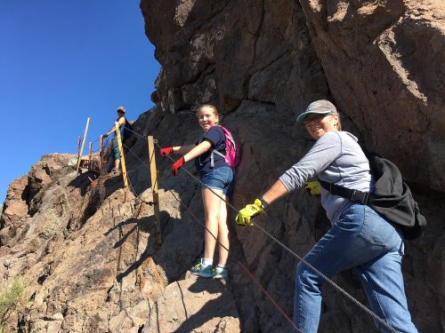 Two people climbing a rocky trail, using ropes for support, with a third person visible in the background. Clear blue sky.