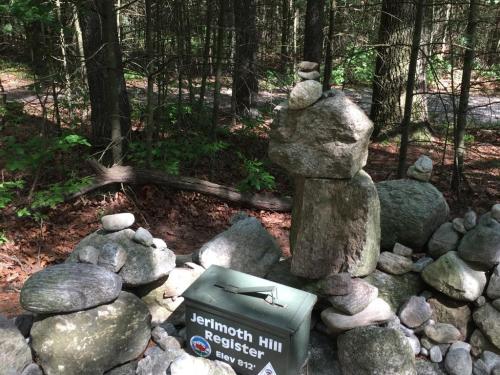 A stone structure with stacked rocks beside a register box in a wooded area. Green foliage surrounds the scene.