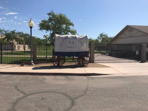 A covered wagon labeled "U.S. Army Transport" parked near a fence and building on a sunny day.