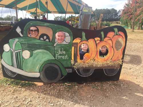 A family poses behind a painted cutout of a vintage truck with pumpkins, smiling at a fall festival.