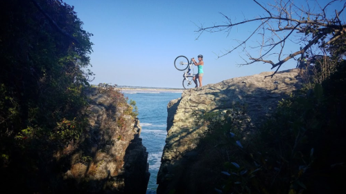 A person stands on a rocky ledge by the water, holding a bicycle above their head against a clear blue sky.