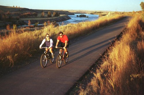 Two cyclists ride along a scenic path beside a river, surrounded by tall grass and autumn foliage.