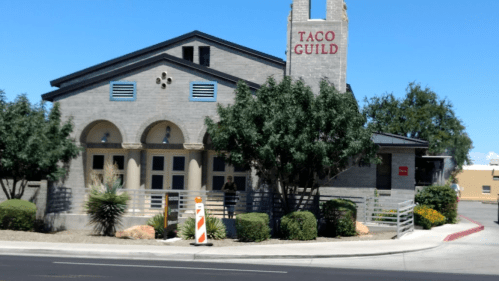 A building with a sign reading "Taco Guild," featuring a modern design and surrounded by trees and landscaping.