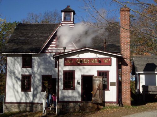 A quaint cider mill with a chimney emitting steam, surrounded by trees and a clear blue sky.