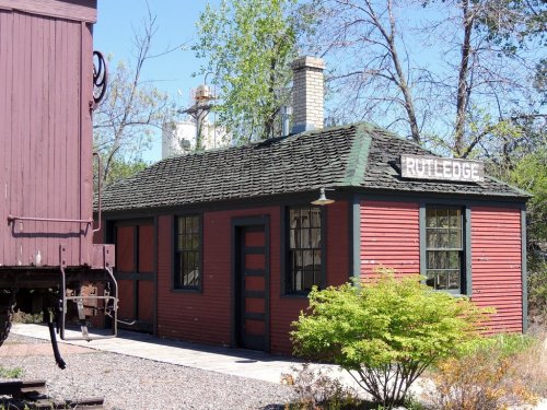 A small red building with a green door and windows, labeled "Rutledge," next to a vintage train car.