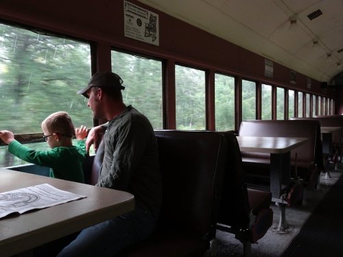 A man and a child sit in a train car, looking out the window at passing scenery. The interior is empty and rustic.