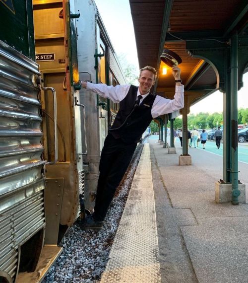 A man in a suit leans out of a vintage train, smiling, with a platform and trees in the background.