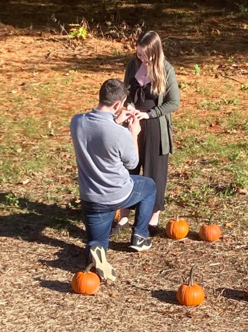 A man kneels to propose to a woman surrounded by small pumpkins in a natural setting.