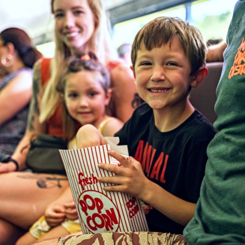 A smiling boy holds a popcorn bucket, sitting with a girl and adults in a lively setting.