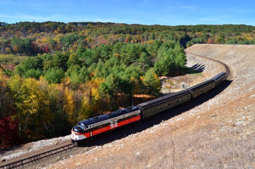 A vintage train winds through a colorful autumn landscape with trees and hills in the background.