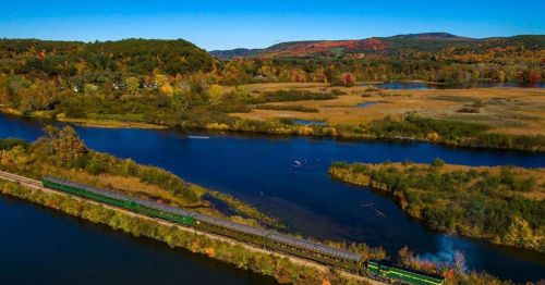 A scenic view of a train traveling along a river, surrounded by vibrant autumn foliage and rolling hills.