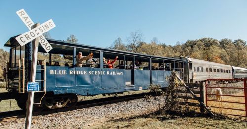 A vintage train on the Blue Ridge Scenic Railway, with passengers waving from an open car near a railroad crossing sign.