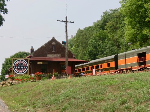 Historic railway station with a vintage train, surrounded by greenery and flowers, featuring a prominent railway sign.