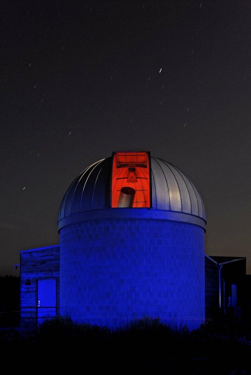 A dome observatory at night, illuminated in blue and red, with stars streaking across the dark sky.
