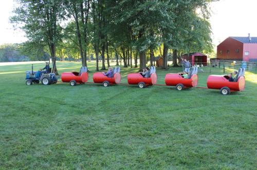 A tractor pulls a train of red barrel-shaped carts on a grassy field, surrounded by trees and a red barn in the background.