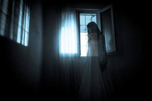 A ghostly figure in a white dress stands by a window, illuminated by soft light in a dark room.