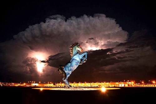 A striking blue horse statue rears against a dramatic night sky with lightning and clouds in the background.