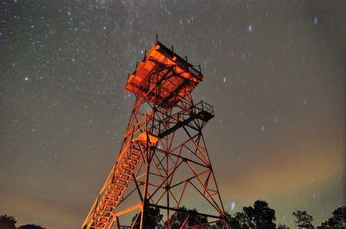 A tall fire tower illuminated against a starry night sky, with trees in the foreground and streaks of stars above.