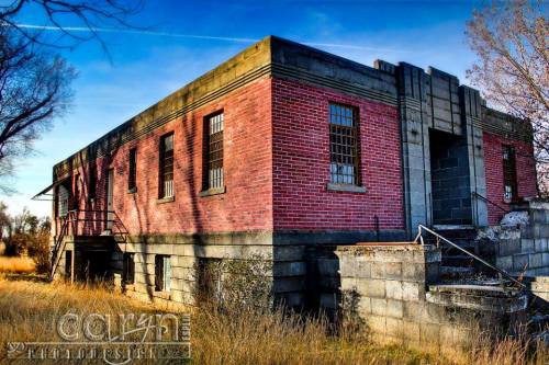 Abandoned brick building with large windows, set in a grassy area under a clear blue sky.